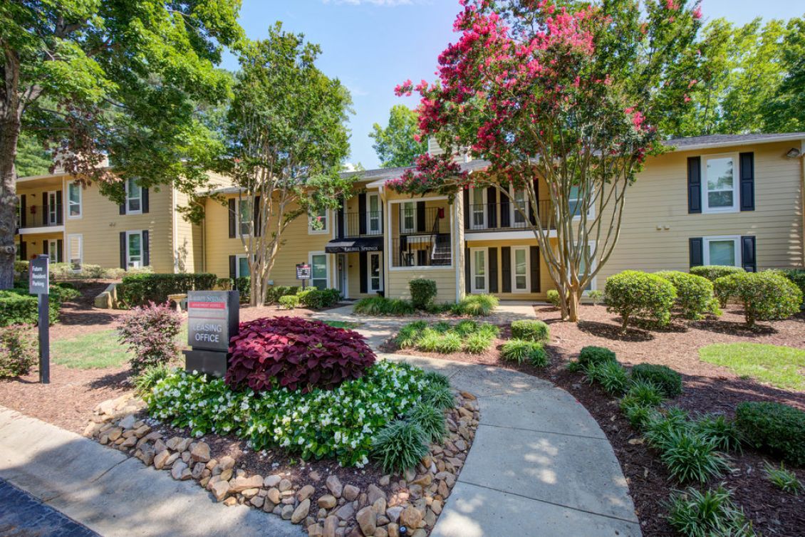 Pathway leading to a leasing office in front of a tan apartment building with trees and landscaped bushes.