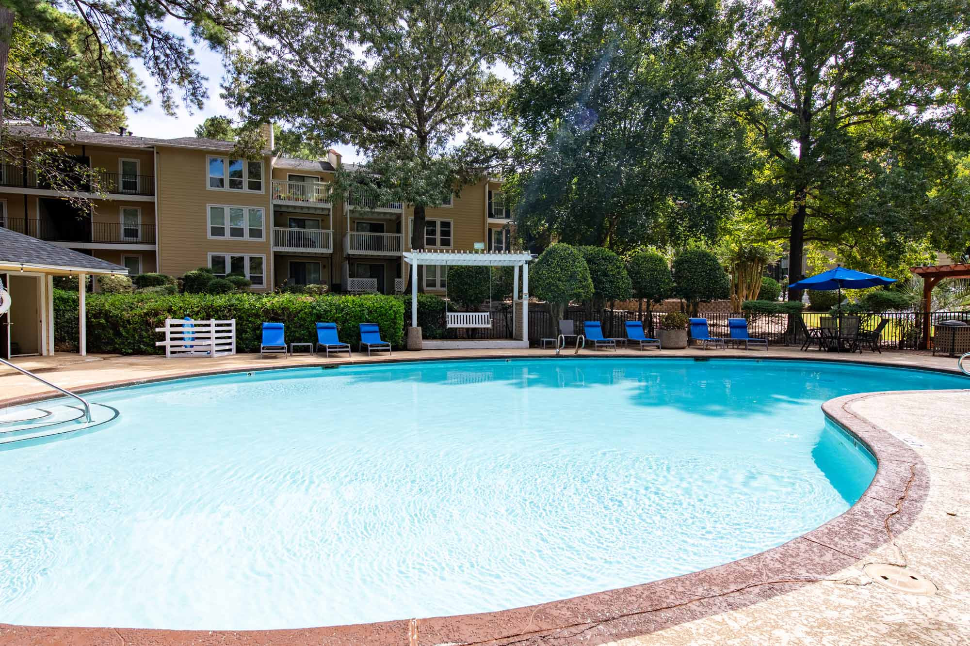 Outdoor swimming pool surrounded by lounge chairs, greenery, and apartment buildings on a sunny day.
