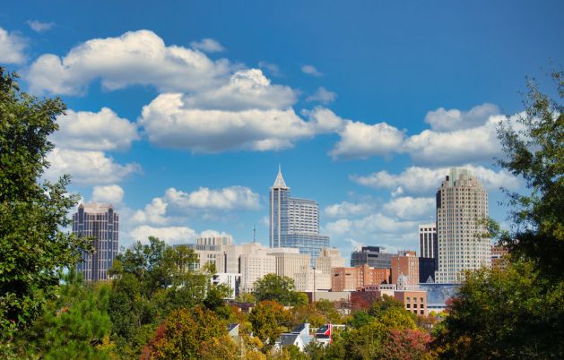 Downtown city skyline with modern buildings, trees in the foreground, and a blue sky with scattered clouds.