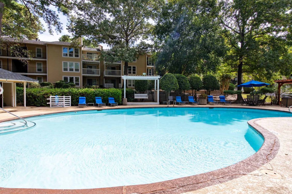 Outdoor swimming pool surrounded by lounge chairs, greenery, and apartment buildings on a sunny day.