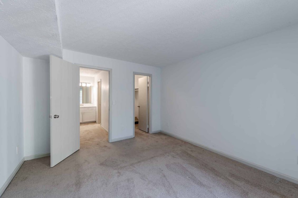 Empty bedroom with beige carpet, white walls, open door to bathroom, and a small walk-in closet.