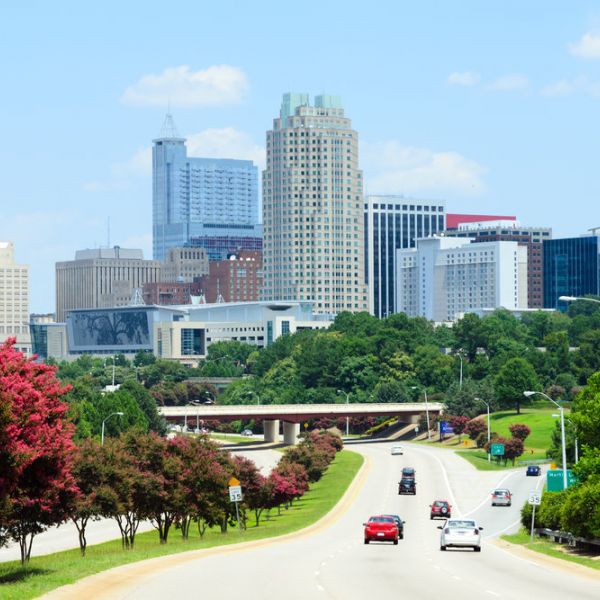 Cars drive toward the downtown Raleigh, North Carolina skyline under a blue sky with green trees and flowers.