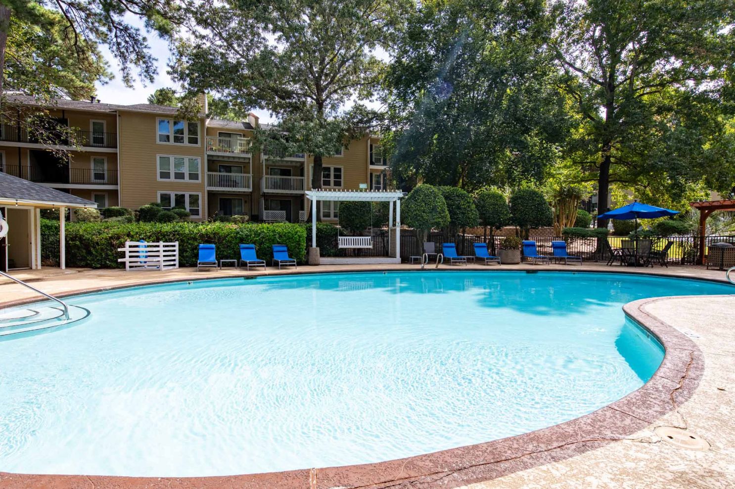 Outdoor swimming pool surrounded by lounge chairs, greenery, and apartment buildings on a sunny day.