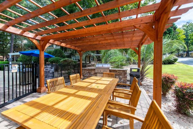 Wooden outdoor dining table and chairs under a pergola near a grill, with trees and a pool in the background.