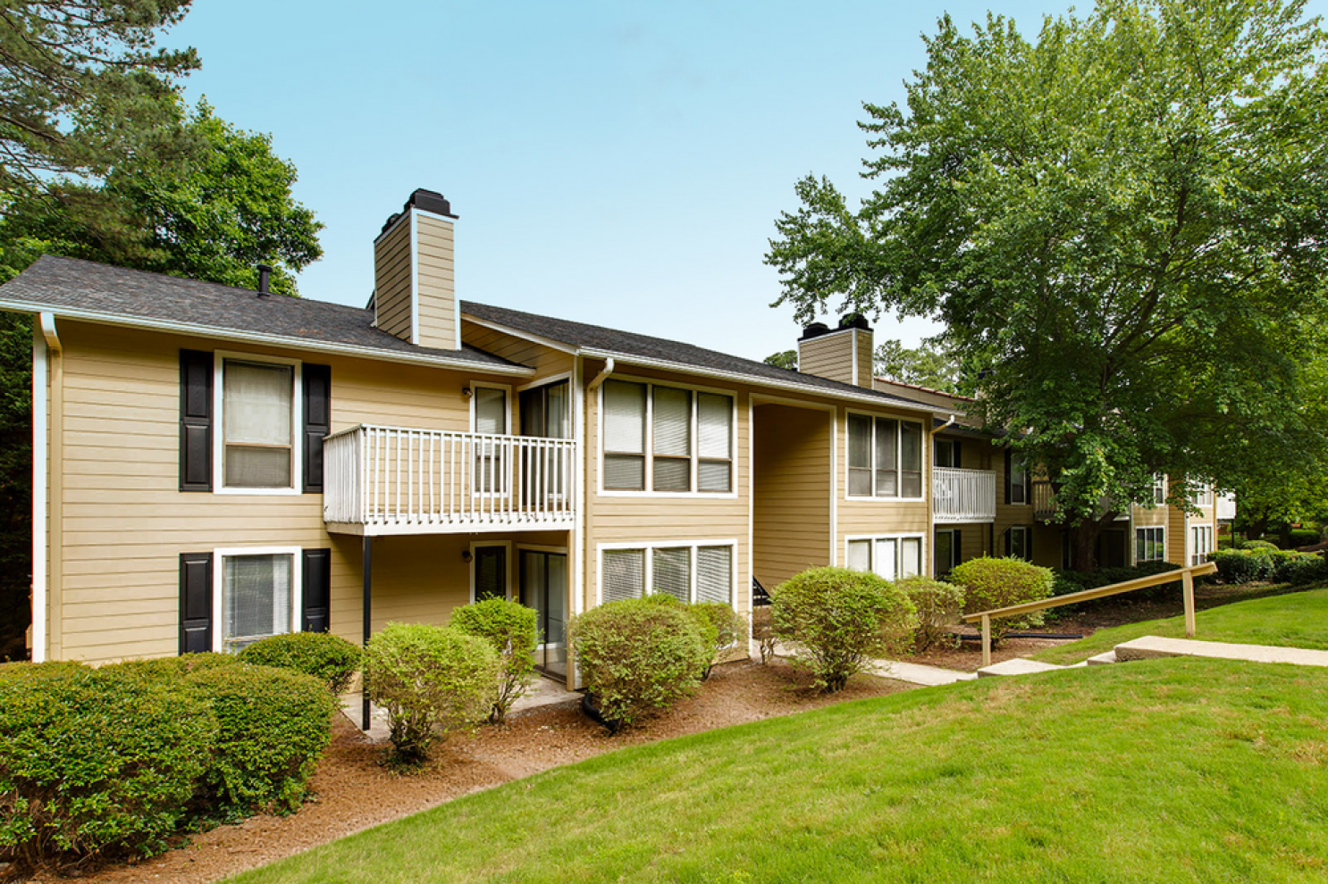 Tan two-story apartment building with balconies, surrounded by green trees, bushes, and a grassy lawn.