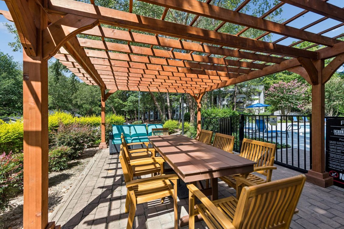 Wooden pergola covering a long outdoor dining table with chairs next to a pool fence.