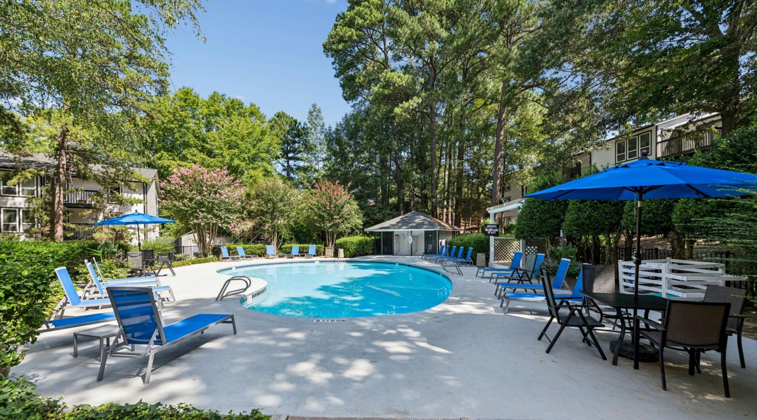 An outdoor swimming pool surrounded by lounge chairs, patio dining sets, and tall trees.