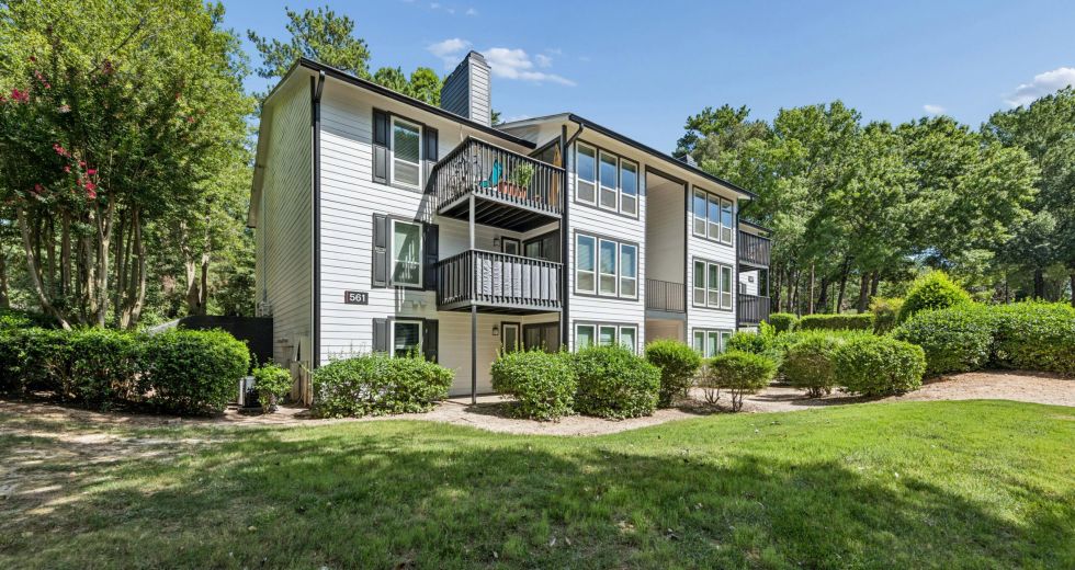 A three-story white apartment building with black shutters and balconies surrounded by trees and a lawn.