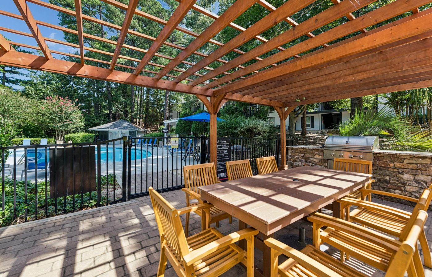 Outdoor patio with a wooden pergola, dining table, chairs, and built-in grill next to a fenced swimming pool area.