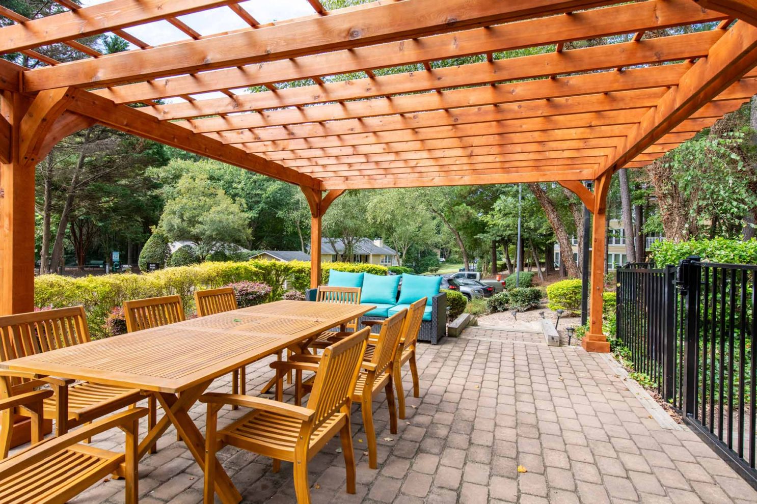 Wooden pergola over outdoor dining set with cushions on a brick patio, surrounded by greenery.