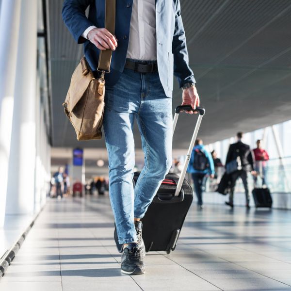 Person walking in an airport terminal with a suitcase and shoulder bag, other travelers in background.