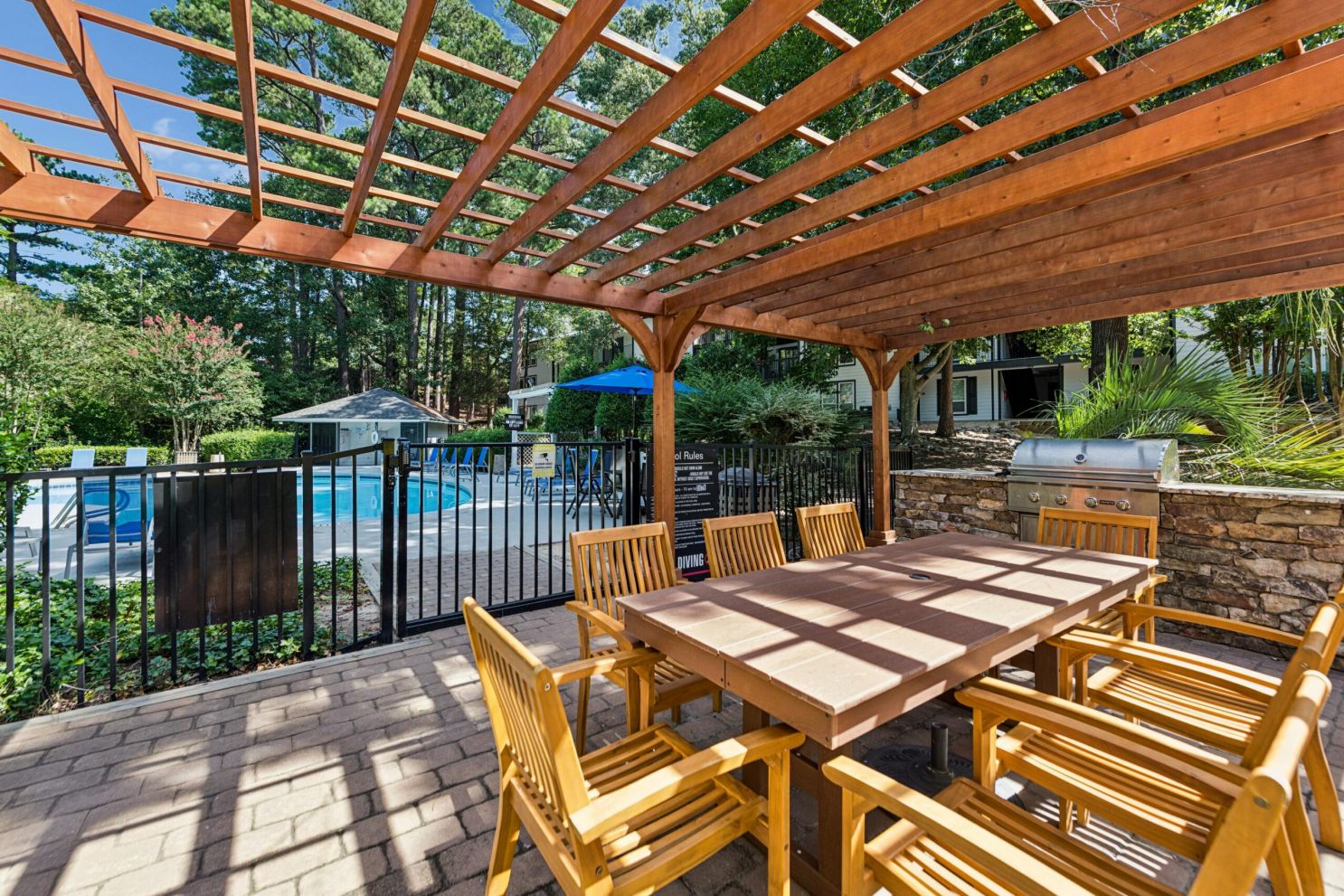 Outdoor patio with a wooden pergola, dining table, chairs, and built-in grill next to a fenced swimming pool area.